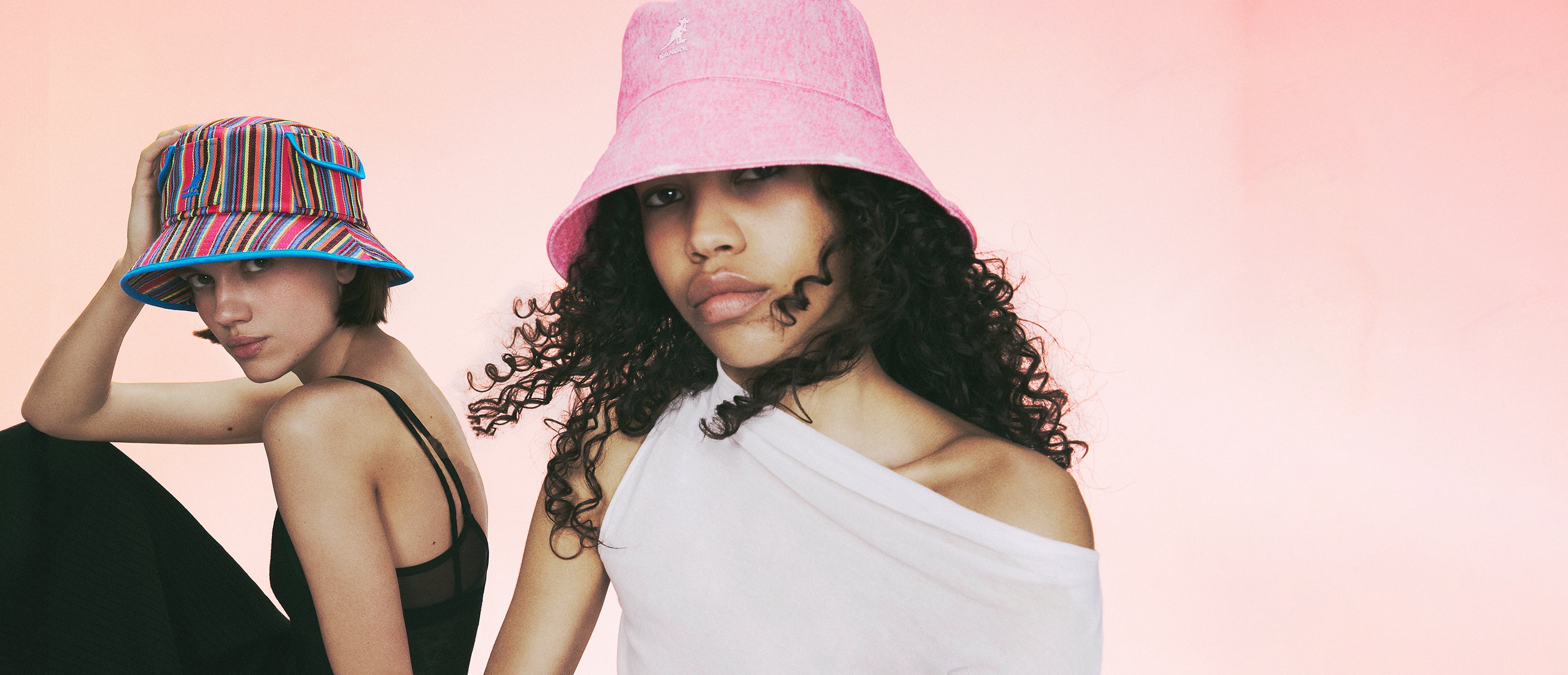 Two women wearing Kangol bucket hats against a pink gradient background—one in a vibrant multicolored striped hat with blue trim and black strappy outfit, the other in a solid pink bucket hat and white off-shoulder top.