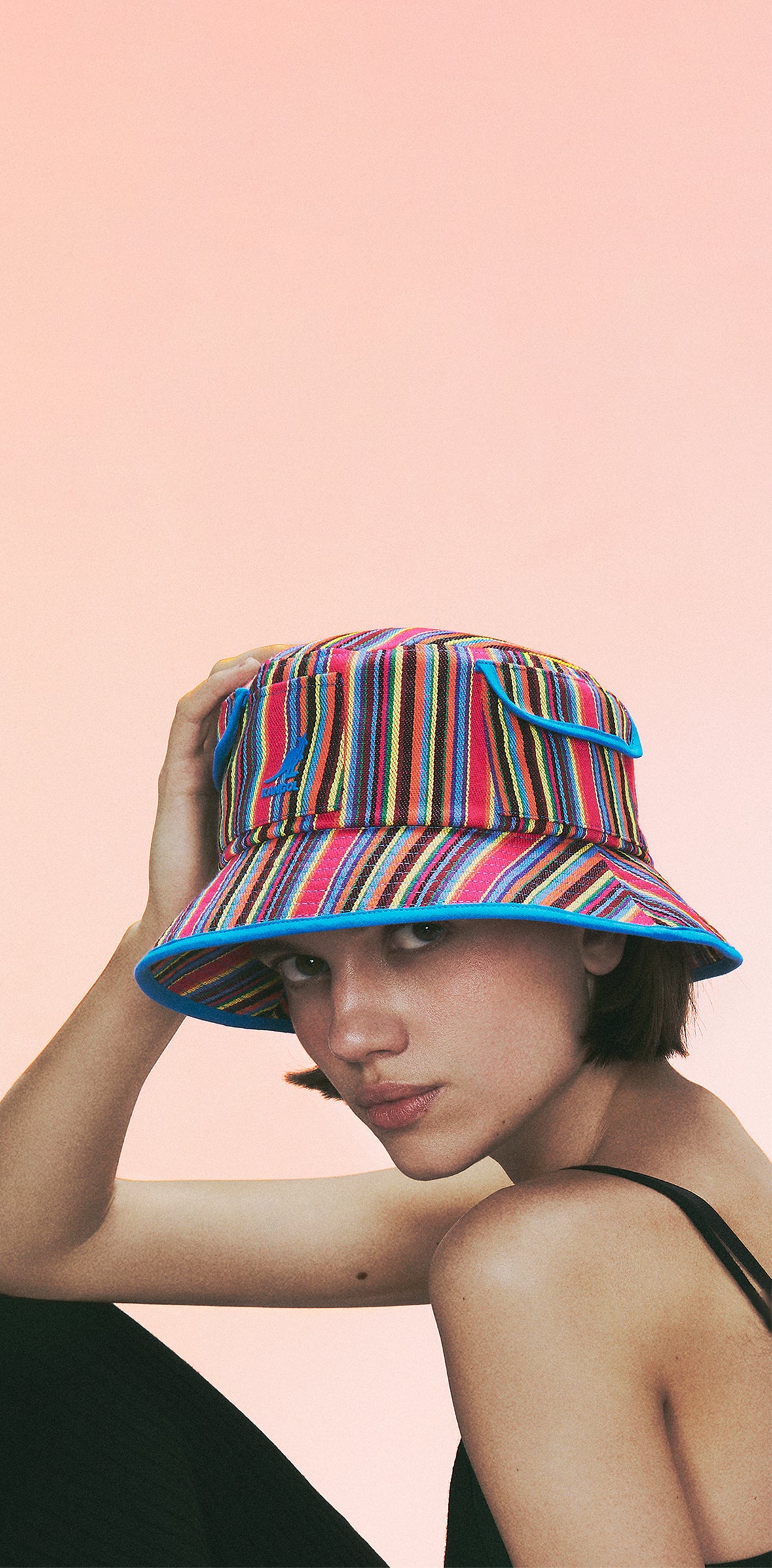 Woman wearing a multicolor striped Kangol bucket hat and black dress against a pink gradient background.
