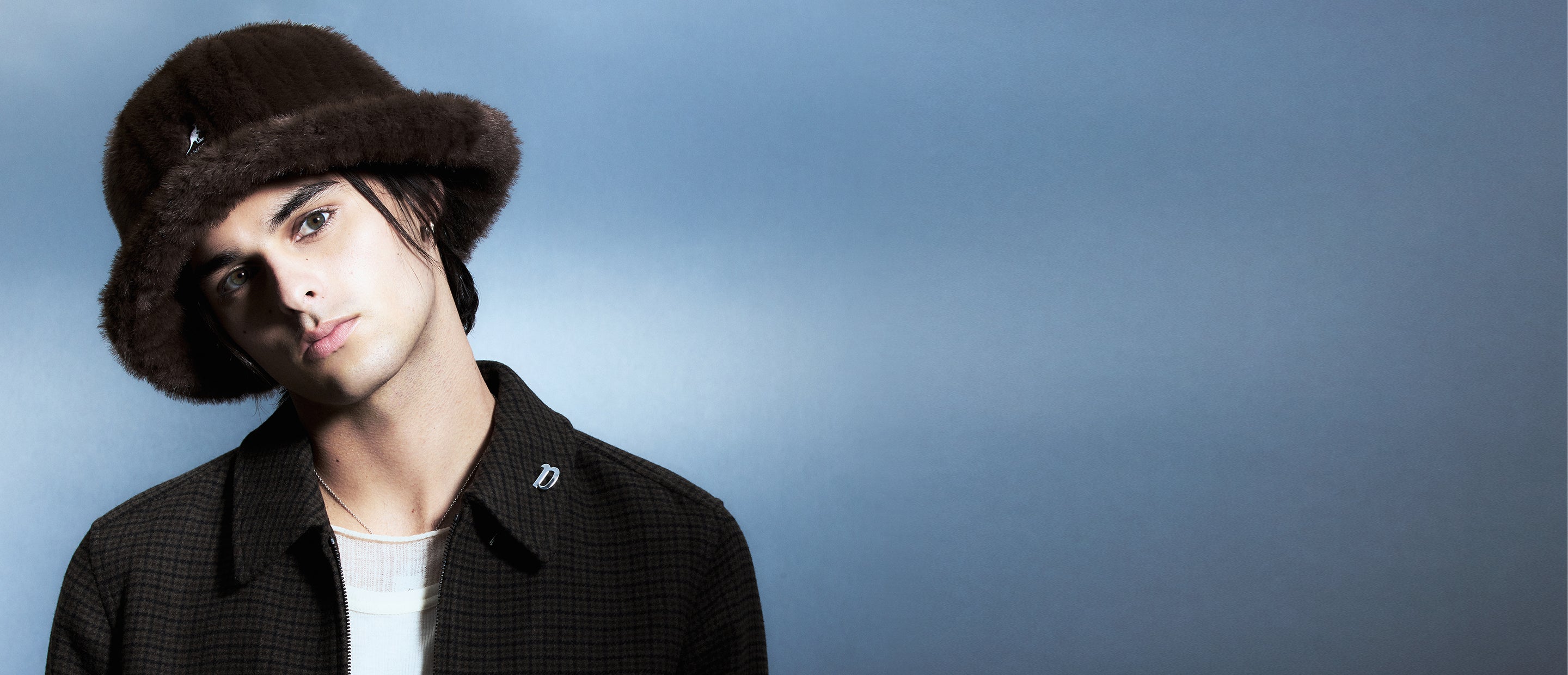 Male model wearing a brown Faux Fur Casual Hat and checked shirt against a blue gradient background.