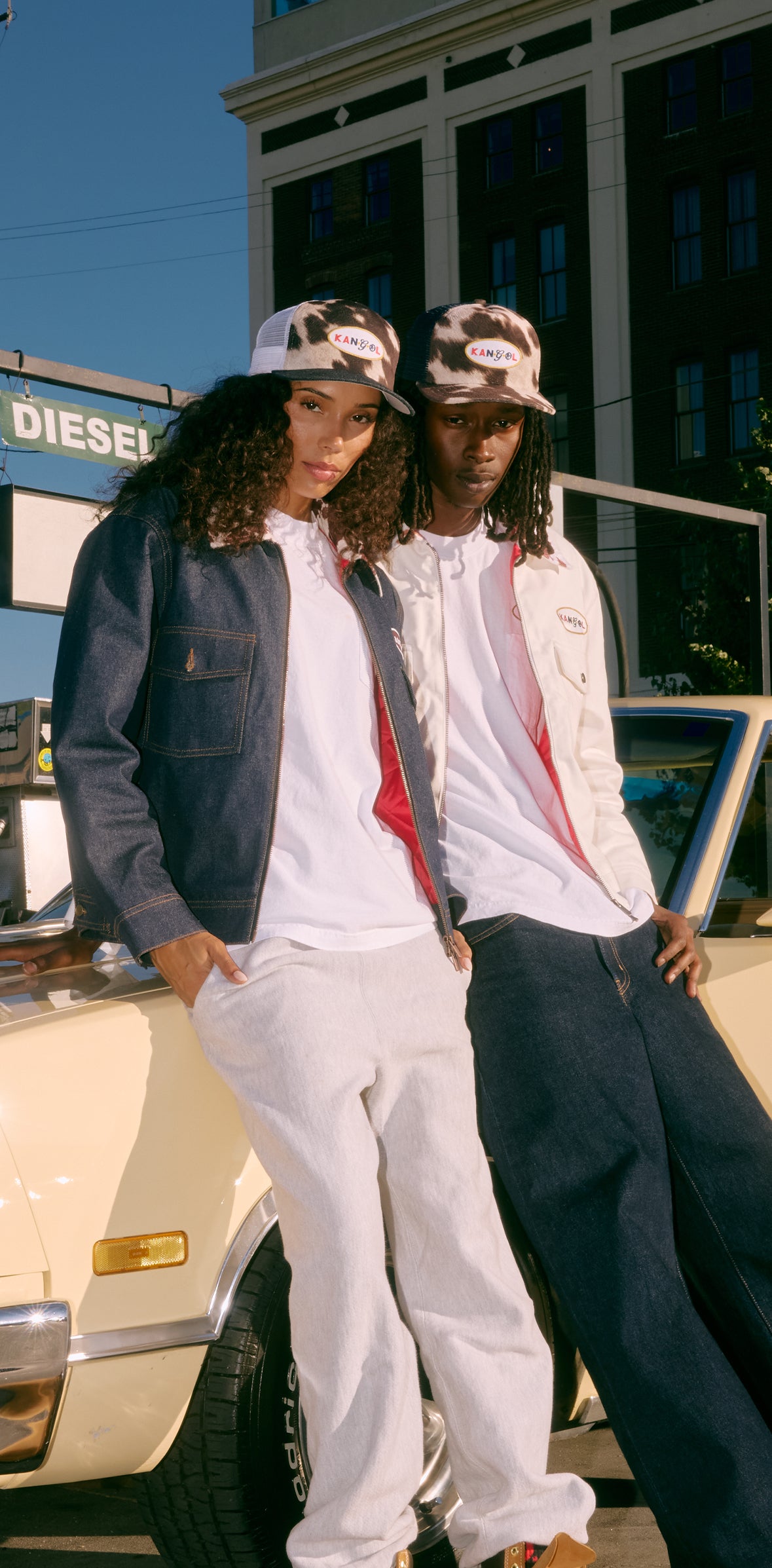 Two people standing next to a car wearing Kangol trucker hats and jackets with a Diesel sign in the background