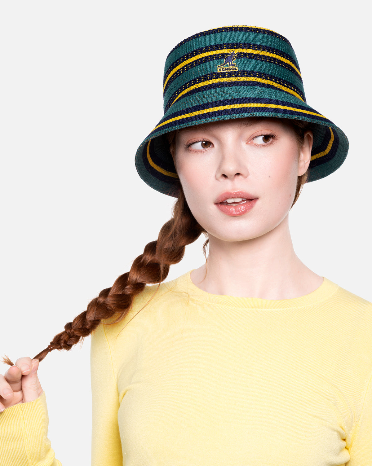 Person wearing a green and yellow striped bucket hat with braided hair on a light gray background