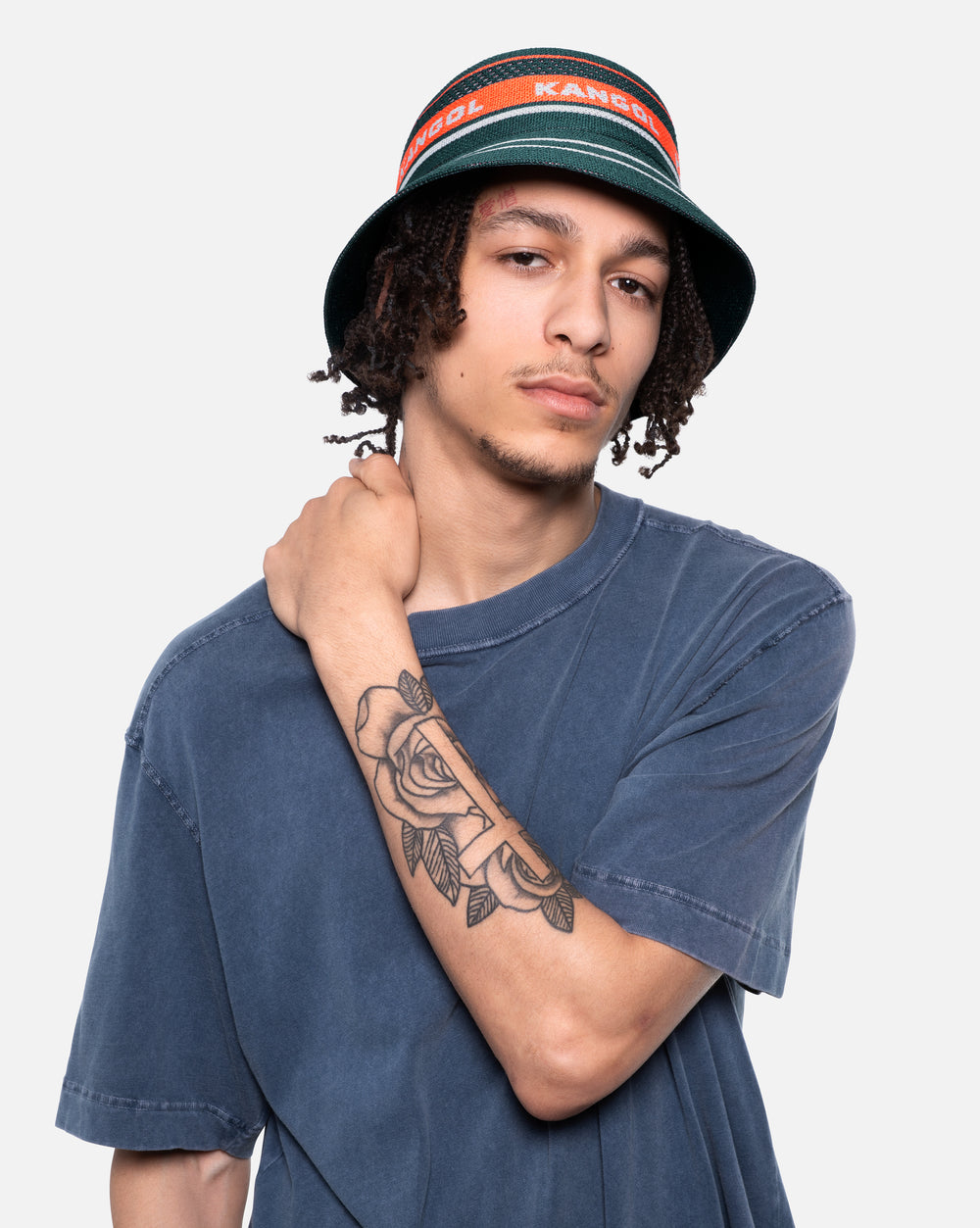 Person wearing a blue t-shirt and a bucket hat with a visible tattoo on the arm, against a plain background