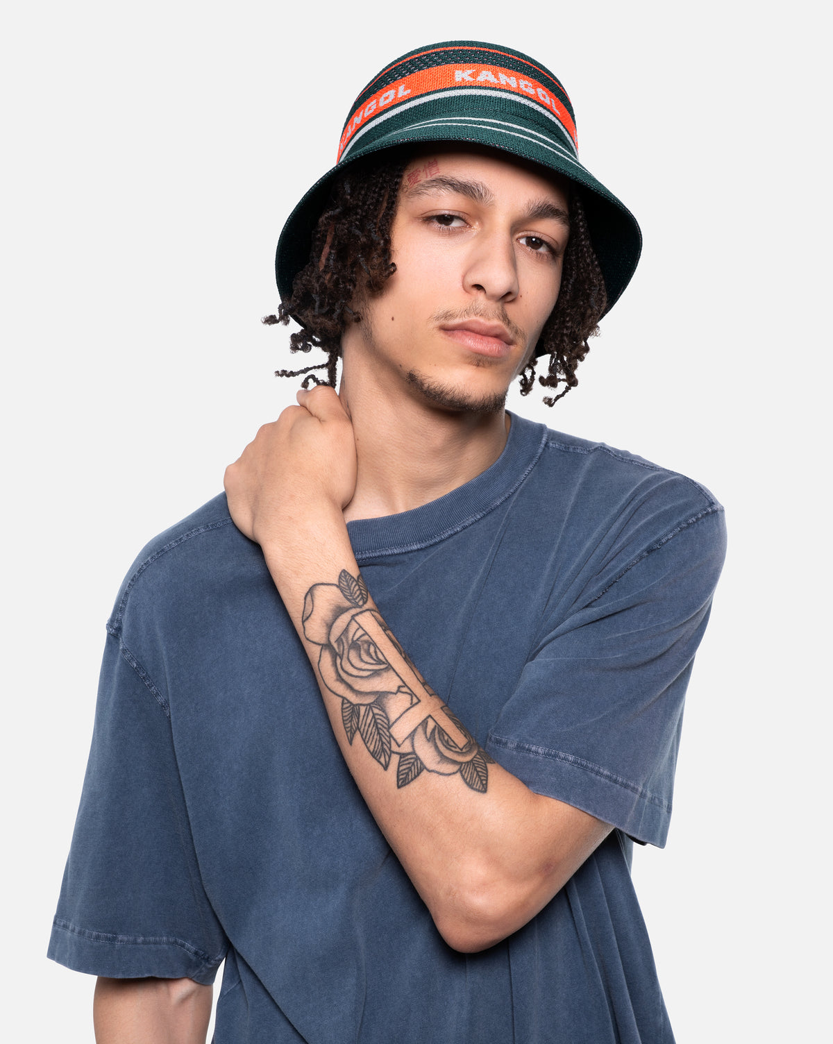 Person wearing a blue t-shirt and a bucket hat with a visible tattoo on the arm, against a plain background