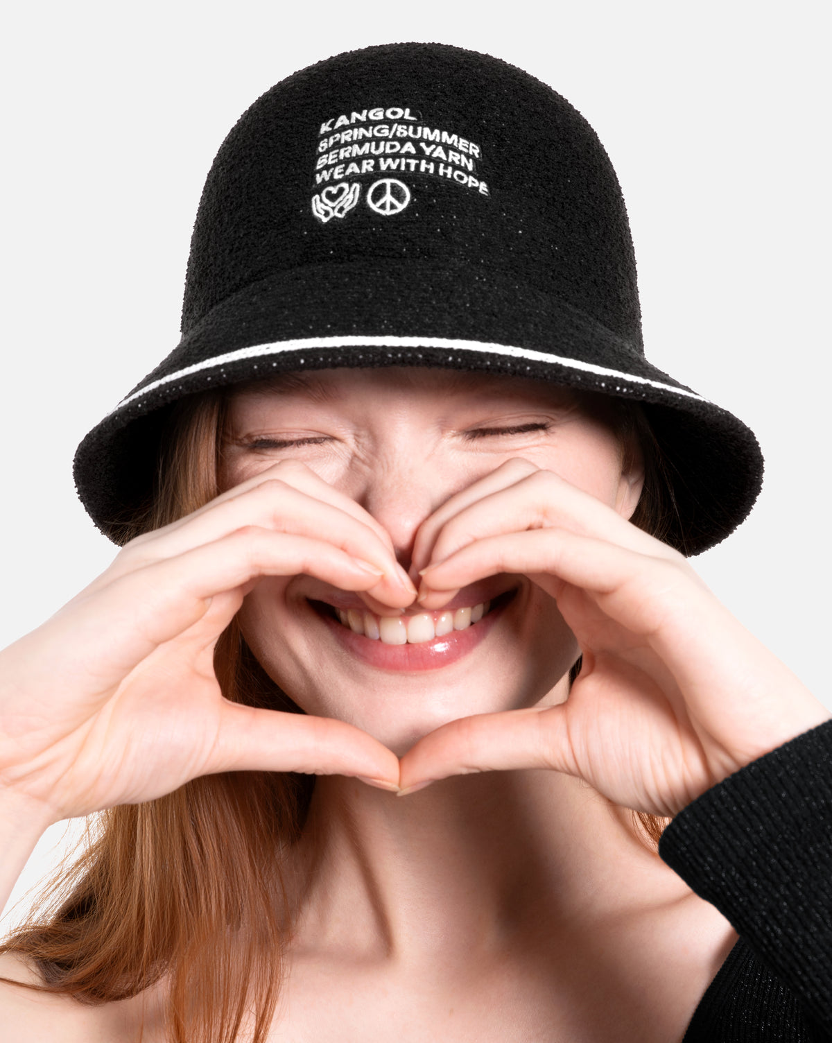 Person wearing a black bucket hat with white text and peace symbol, making a heart shape with their hands.