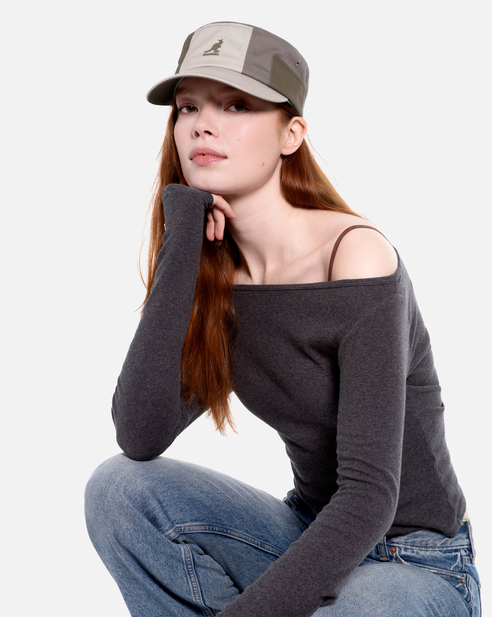 Woman wearing a gray off-shoulder top and jeans with a gray colorblock army cap on a white background