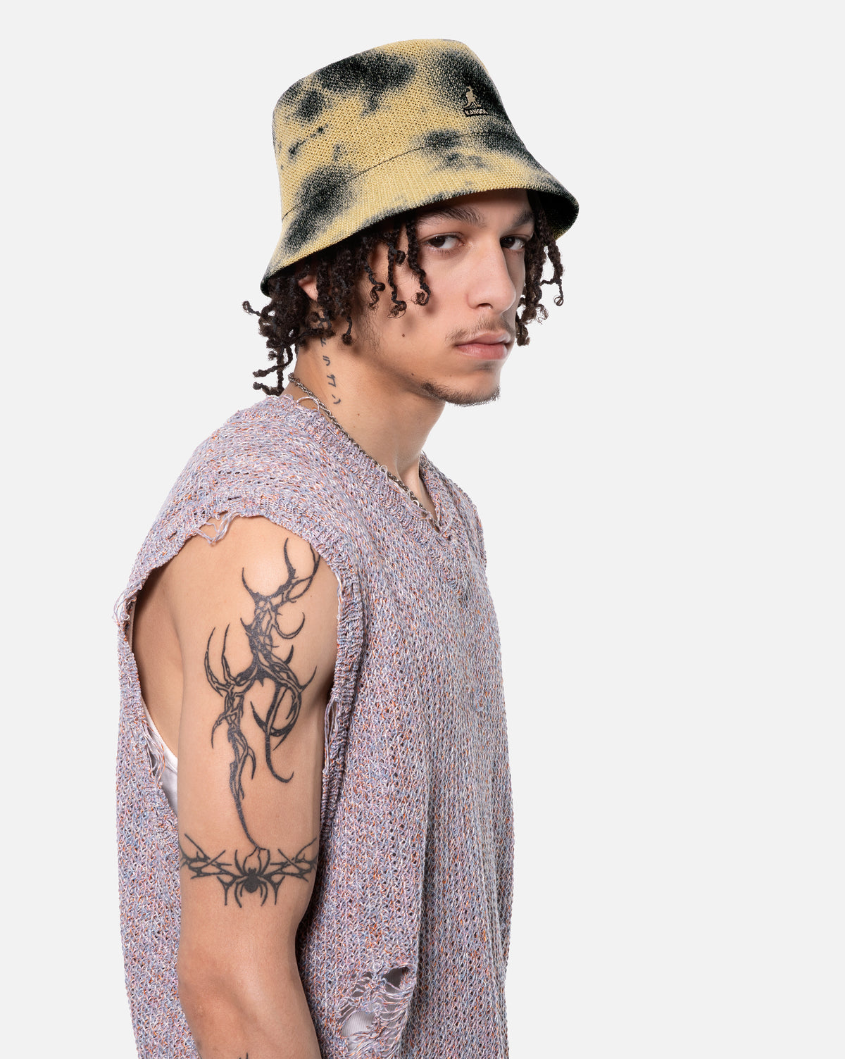 Person wearing a tie-dye bucket hat and sleeveless shirt with tattoos on a white background