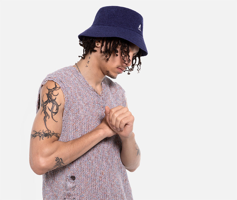 Person wearing a navy bucket hat and sleeveless shirt with tattoos on arms, on a white background