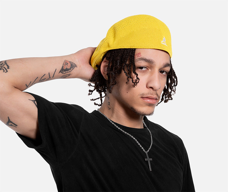 Person wearing a yellow cap and black shirt with tattoos on arms, on a white background
