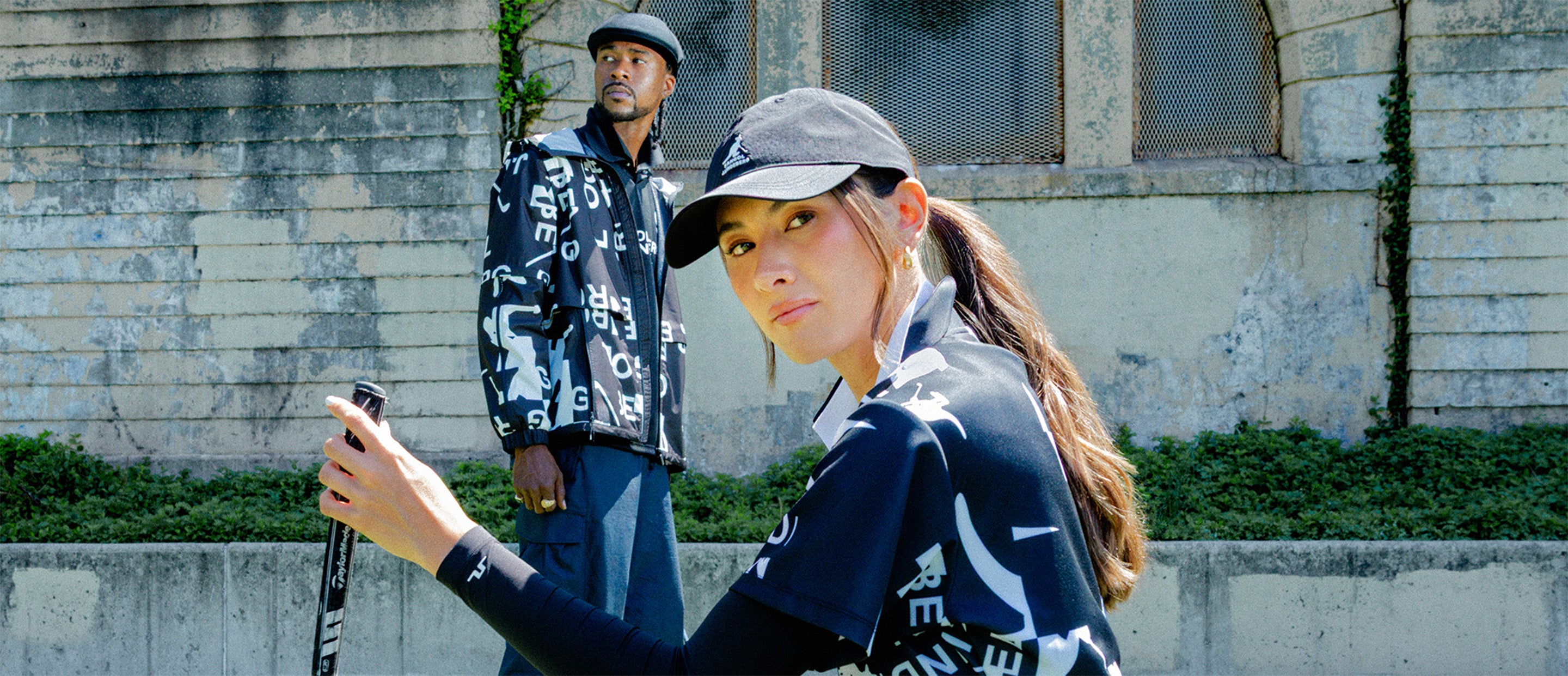 Two models on a sunny turf field wearing black-and-white graphic golf apparel. The woman in the foreground is crouched with a golf club and ball, wearing a black cap, pleated skirt, and white sneakers. The man in the background stands wearing matching patterned outerwear and a flat cap, against a stone wall with arched windows.
