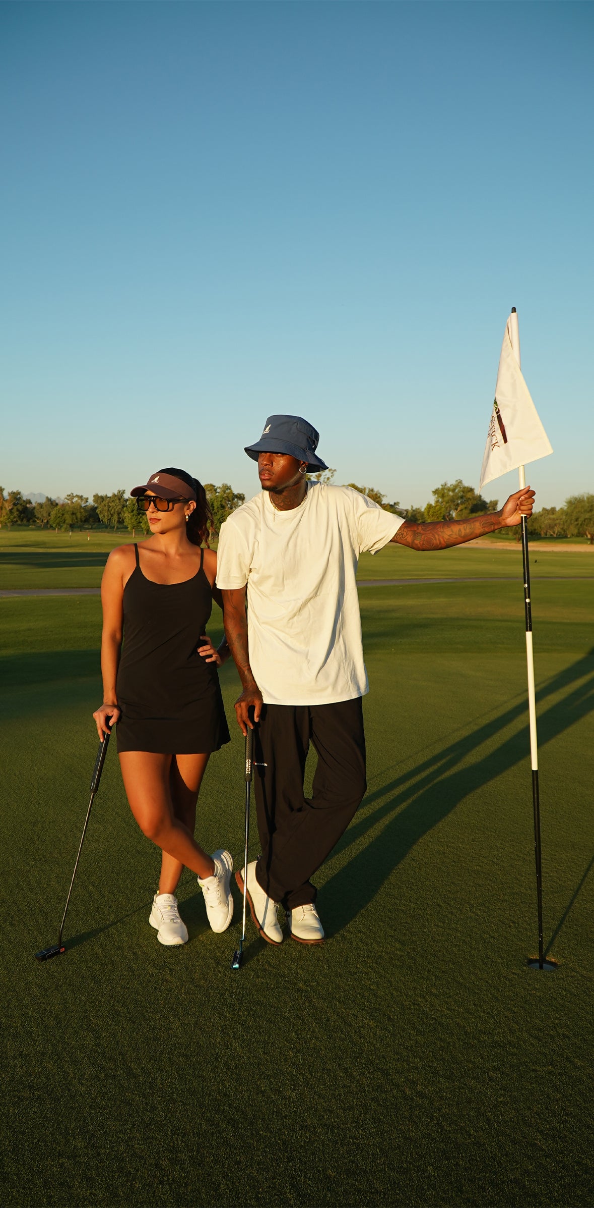 Man and woman standing on golf course holding golf clubs wearing KANGOLF Sport Visor in Burgundy and Kangolf Lahinch Bucket Hat in blue. 